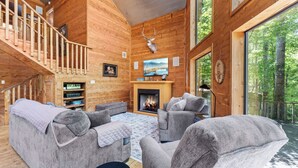 A wooden cabin interior with a fireplace, two grey sofas, a bookshelf, and a deer head mounted on the wall.