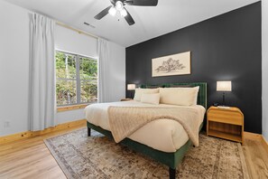 A bedroom with a bed, a nightstand, a ceiling fan, a window with curtains, and a framed picture on the wall.