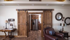 Una habitación con puerta corredera de madera que da a una cocina, un sillón de cuero, una mesita con lámpara y espejos montados en la pared.