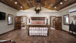 A spacious room with a wooden ceiling, a central bed with pillows, a ceiling fan, and a mounted TV.