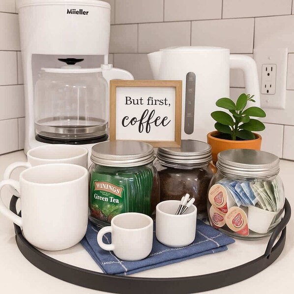 Una encimera de cocina con una cafetera, un cartel que dice 'But first, coffee' y varios objetos relacionados con el café.