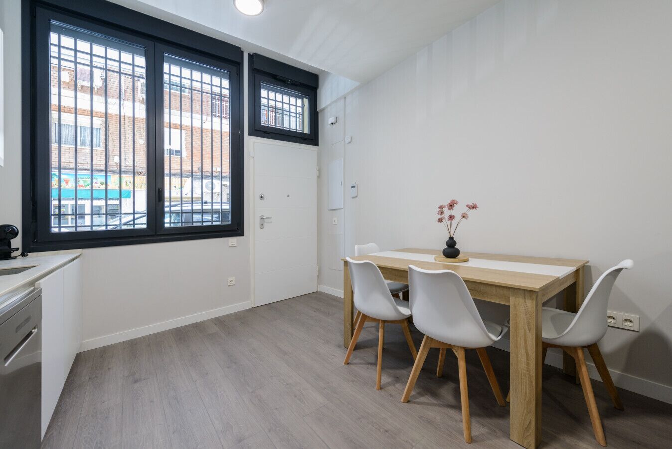 Una habitación con una mesa de madera, cuatro sillas, un jarrón con flores, una ventana con barrotes y una puerta blanca.