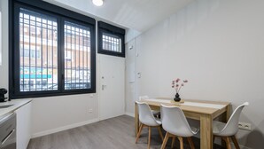 Une pièce avec une table à manger en bois, quatre chaises, un vase de fleurs, une fenêtre avec des barreaux et une porte blanche.