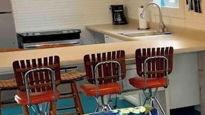 Une cuisine avec un plan de travail blanc, deux suspensions orange, un évier et trois tabourets de bar rouges.