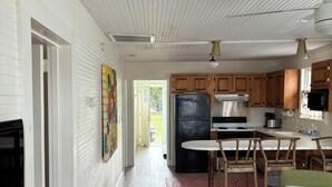 A modern kitchen with wooden cabinets, a black refrigerator, and a dining area with bar stools. - Point Break Historic Beach Cottage (Carolina Beach)