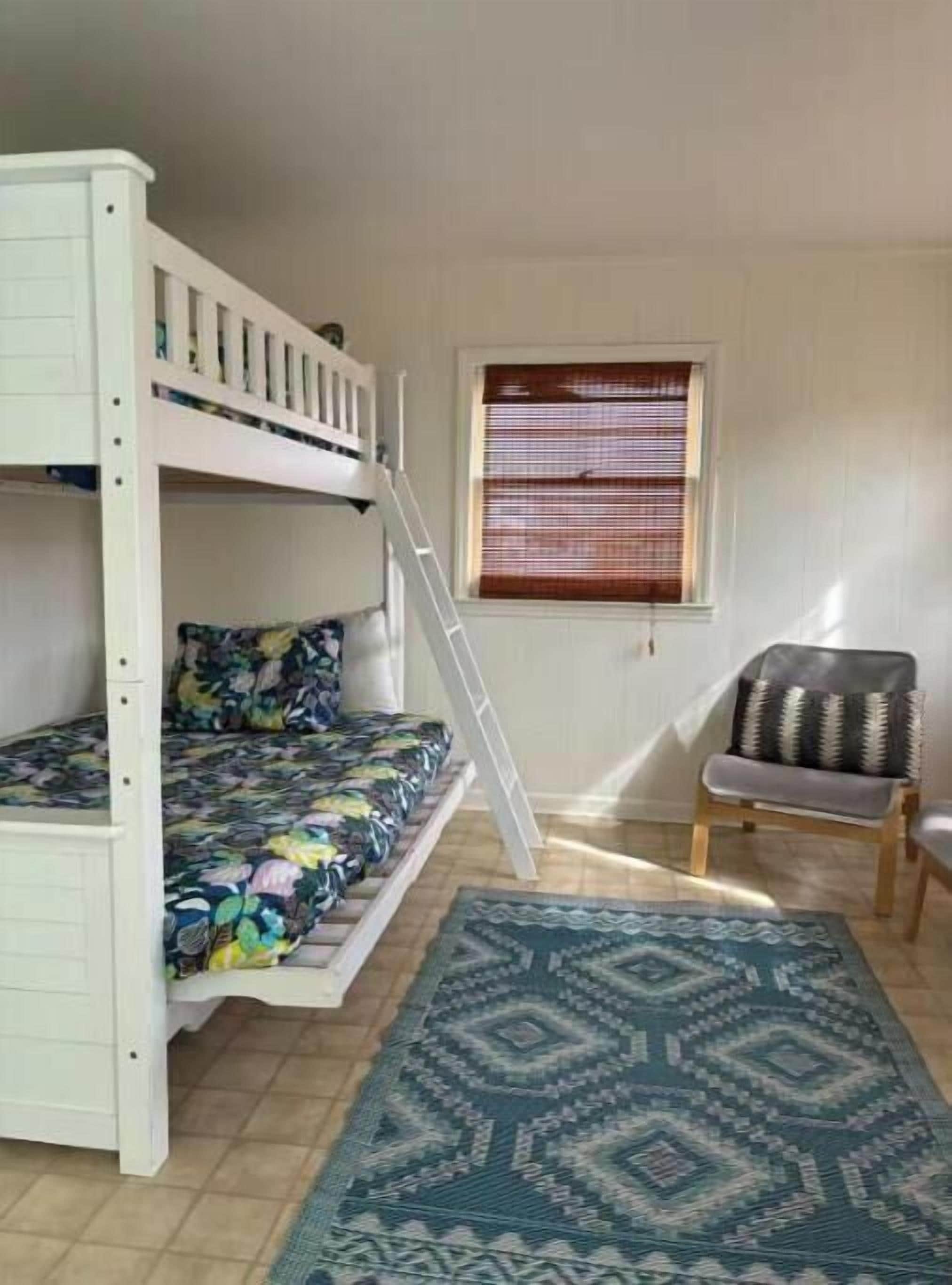 Een stapelbed met een kleurrijk bloemenbedekking, een klein ladder, een raam met zonwering, een stoel met een decoratief kussen en een patroonrijk vloerkleed op een betegelde vloer.