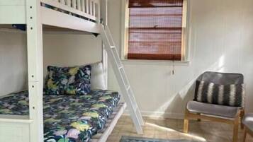 Een stapelbed met een kleurrijk bloemenbedekking, een klein ladder, een raam met zonwering, een stoel met een decoratief kussen en een patroonrijk vloerkleed op een betegelde vloer.