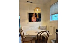 A dining area with a round table, two wooden chairs, a woven pendant light, a bench, and a window with blinds.