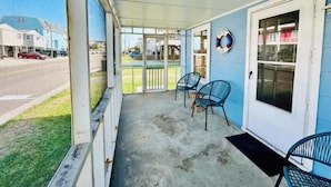 A covered porch with two chairs and a door, overlooking a street with houses and cars.