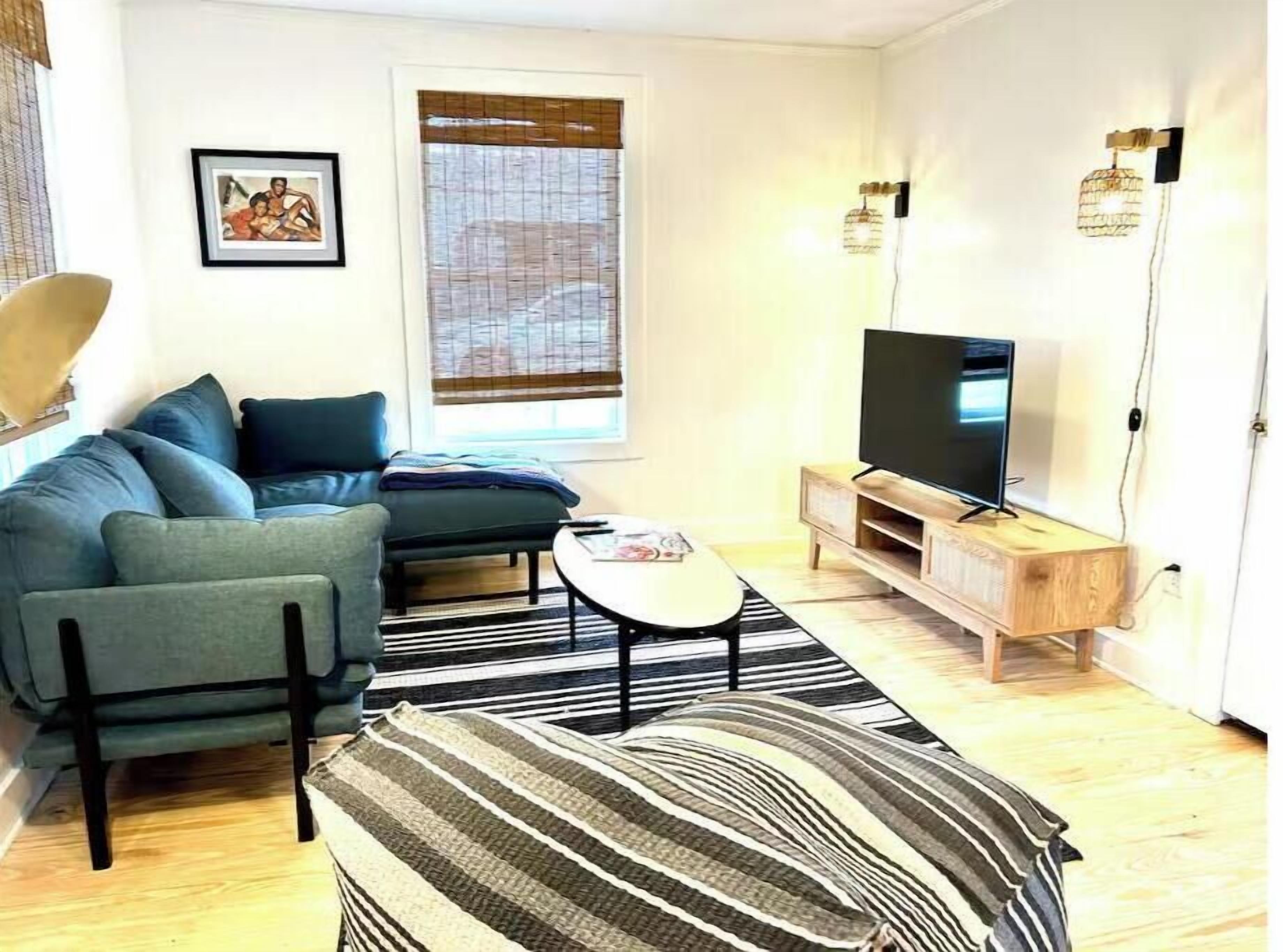 A living room with a sofa, a coffee table, a TV on a wooden stand, a striped rug, and a window with blinds.