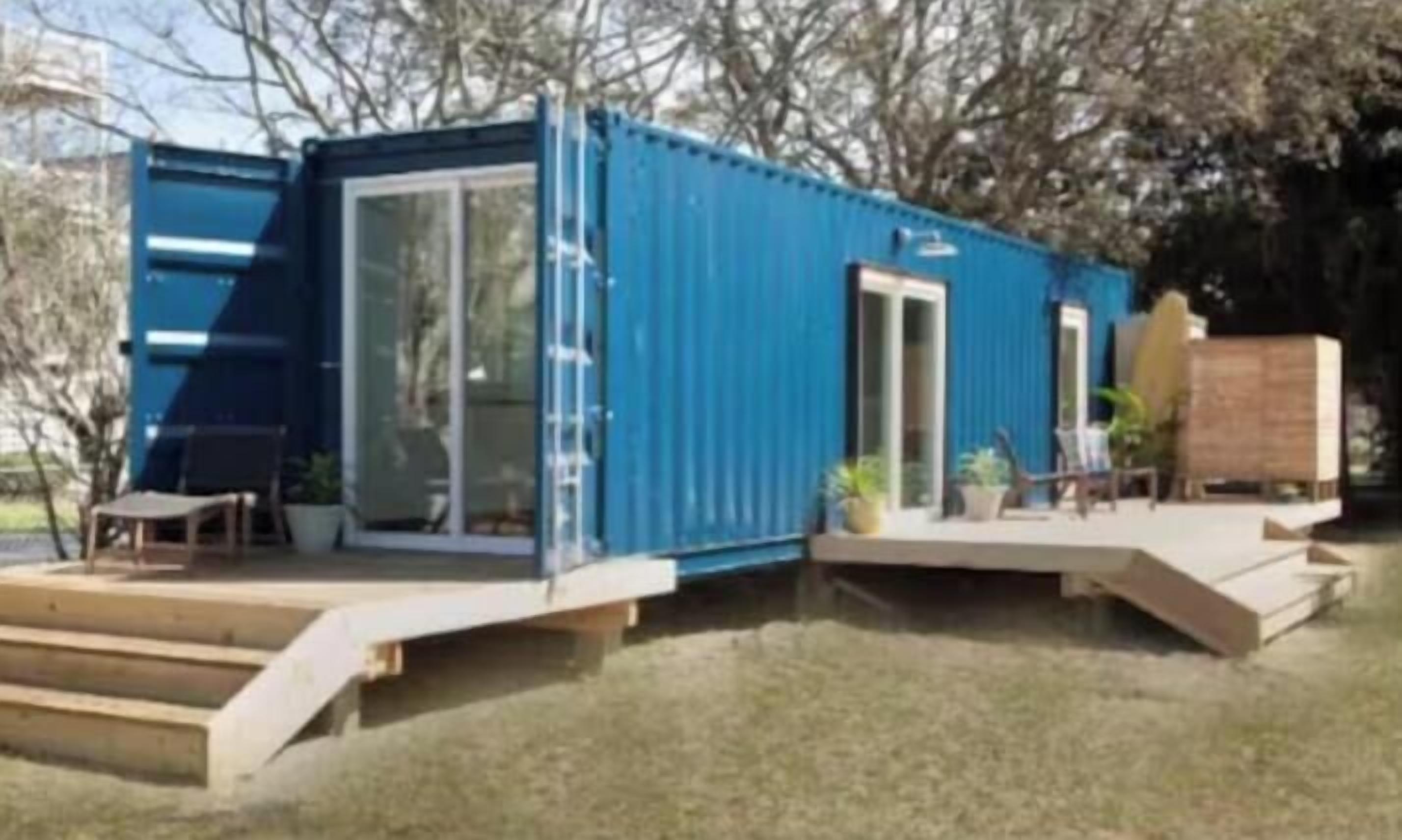 A blue modular building with a glass door, a small outdoor seating area, and a wooden deck.