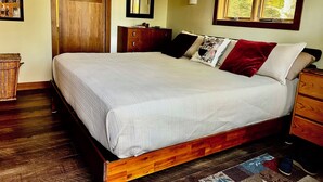 Une chambre à coucher avec un lit en bois, une commode en bois, une table de chevet, un miroir et une fenêtre donnant sur des arbres.