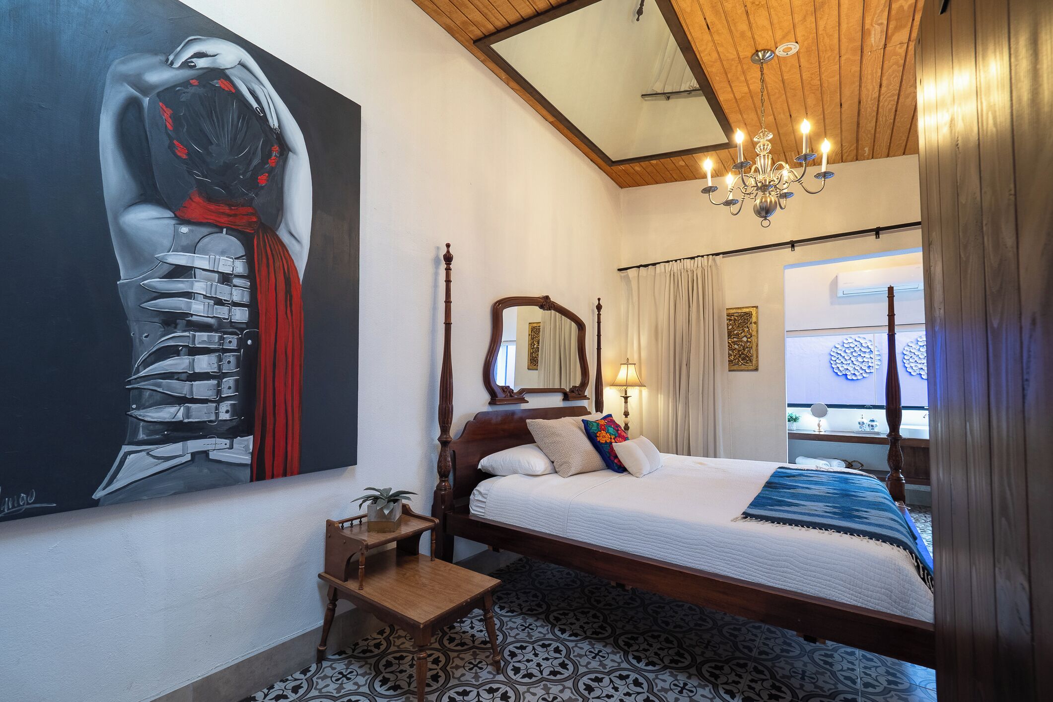 Un dormitorio con una cama grande, cabecera de madera, mesita de noche, espejo, candelabro y un cuadro grande en la pared.