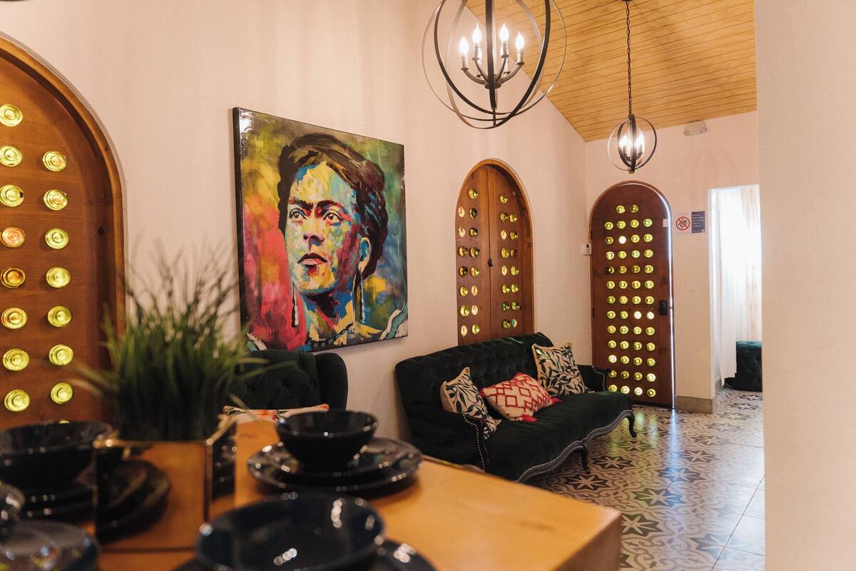 Een kamer met een houten tafel, een zwarte bank en een kleurrijk schilderij aan de muur. Er zijn twee gewelfde deuren met cirkelvormige patronen en een kroonluchter hangt aan het plafond.