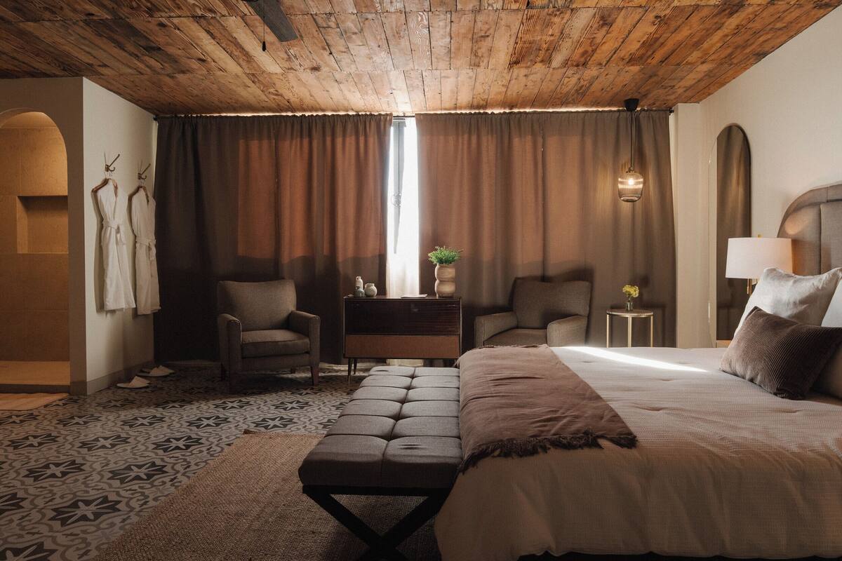 Een slaapkamer met een houten plafond, een groot bed, twee fauteuils, een tafeltje met een plant en een nachtkastje met een lamp.