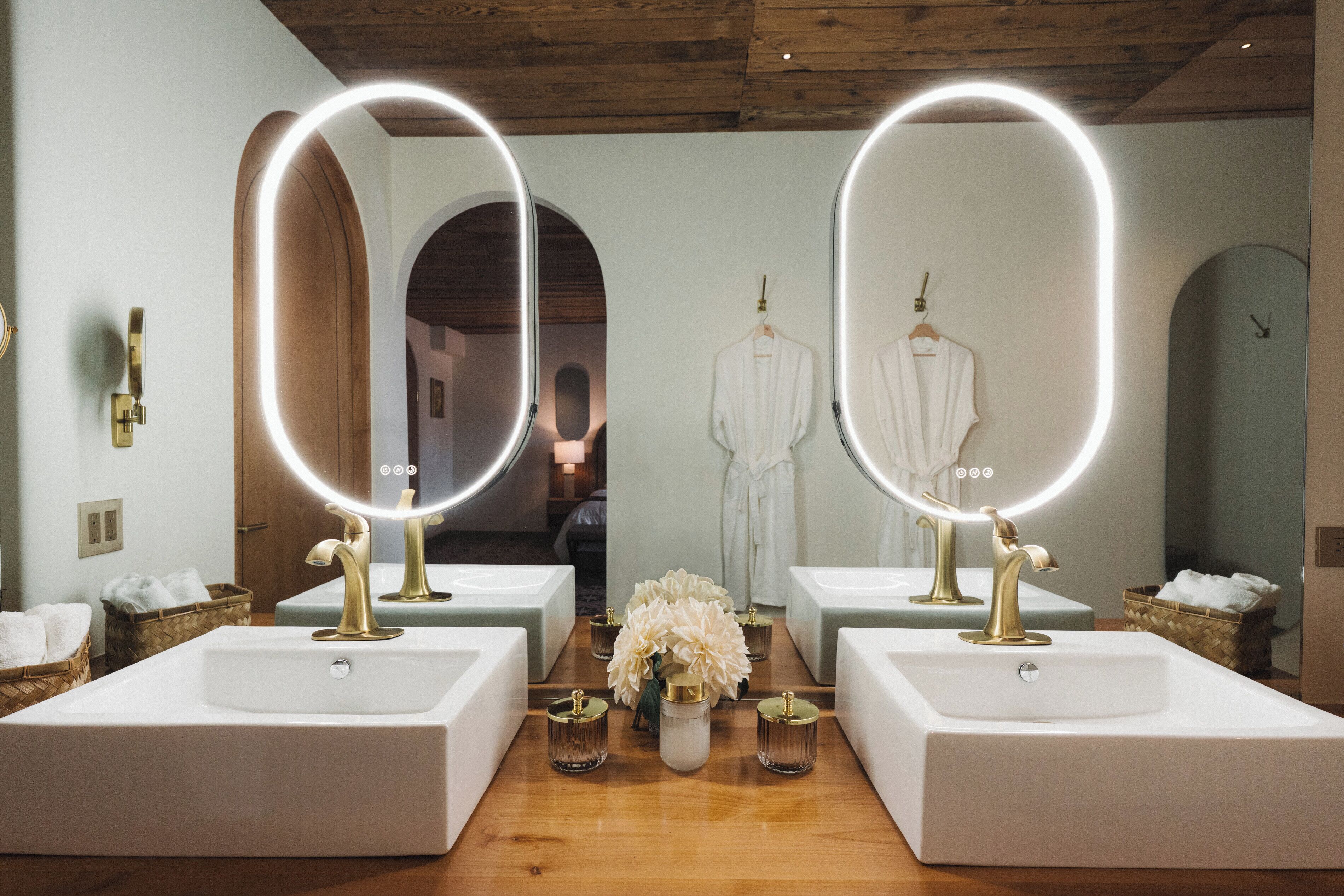 A hotel bathroom with two sinks, each with a mirror and a gold faucet, and a wooden floor.