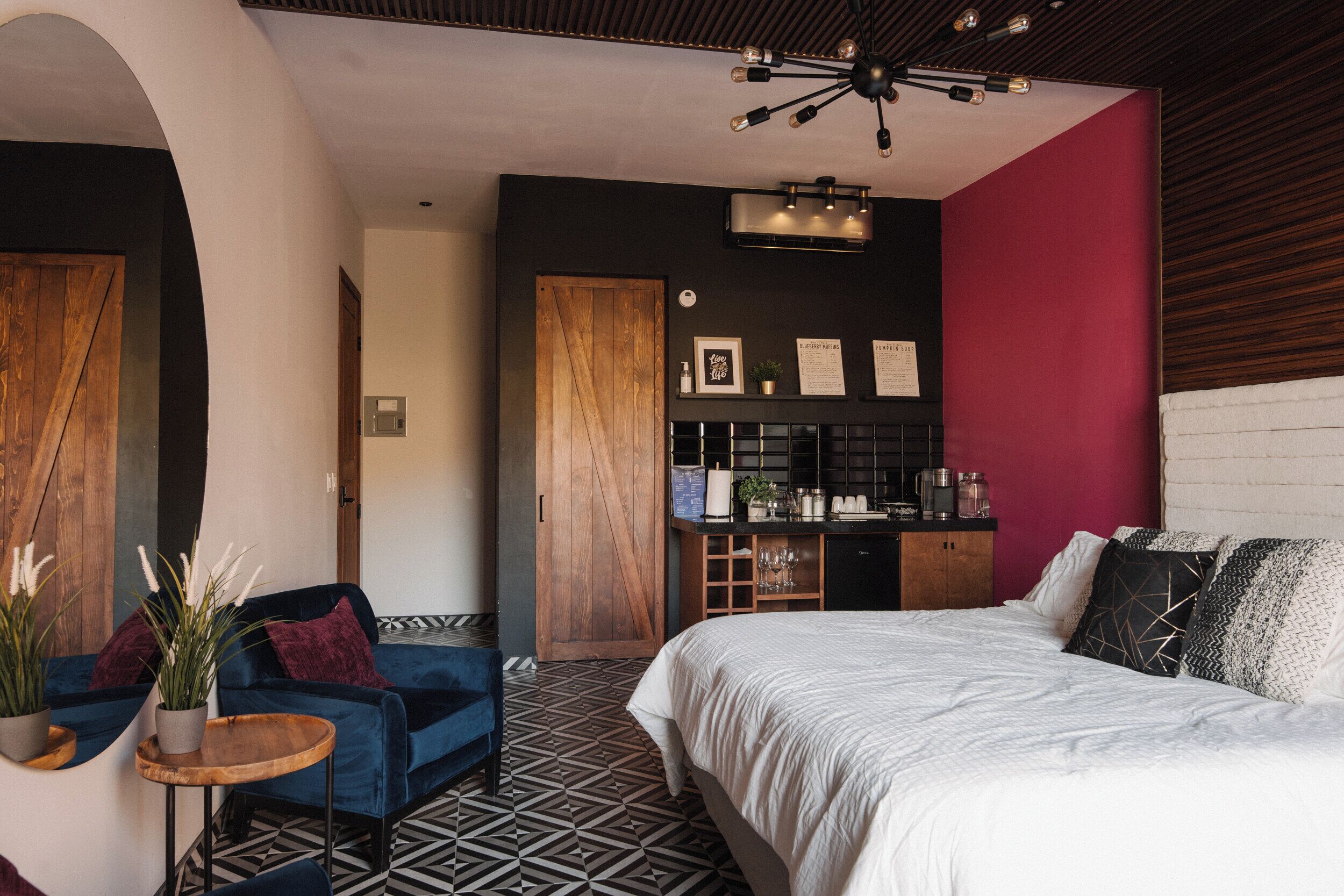 A modern bedroom with a large bed, a blue chair, a small table, and a kitchen area with a red wall.