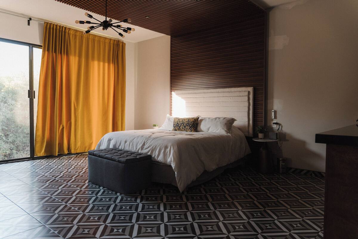Een moderne slaapkamer met een vloer met een geometrisch patroon, een groot bed met hoofdeinde, een nachtkastje en een raam met gele gordijnen.
