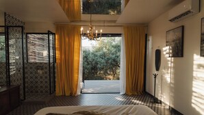 A bedroom with a large bed, a chandelier, a view of trees through a window, and a patterned floor. - Casa Frida (Valle de Guadalupe)