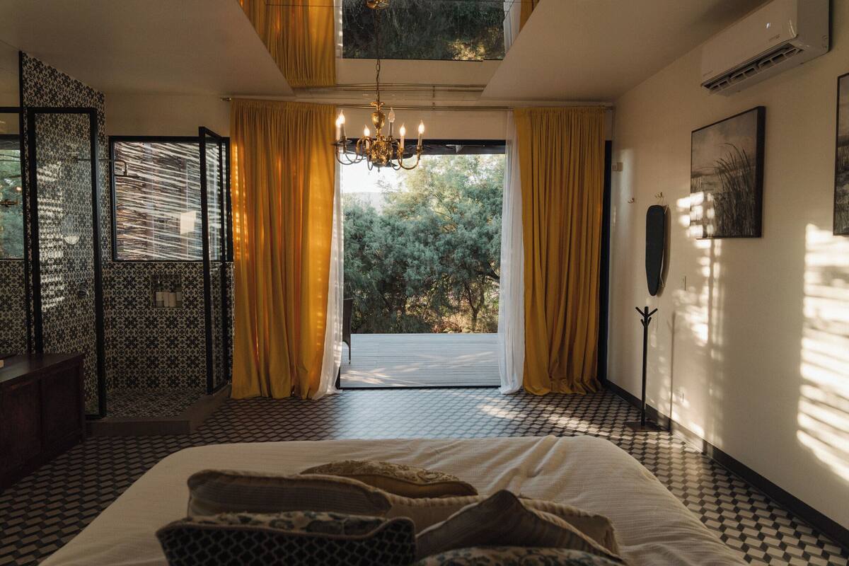 Een slaapkamer met een groot bed, een kroonluchter, uitzicht op bomen door een raam en een vloer met een patroon.