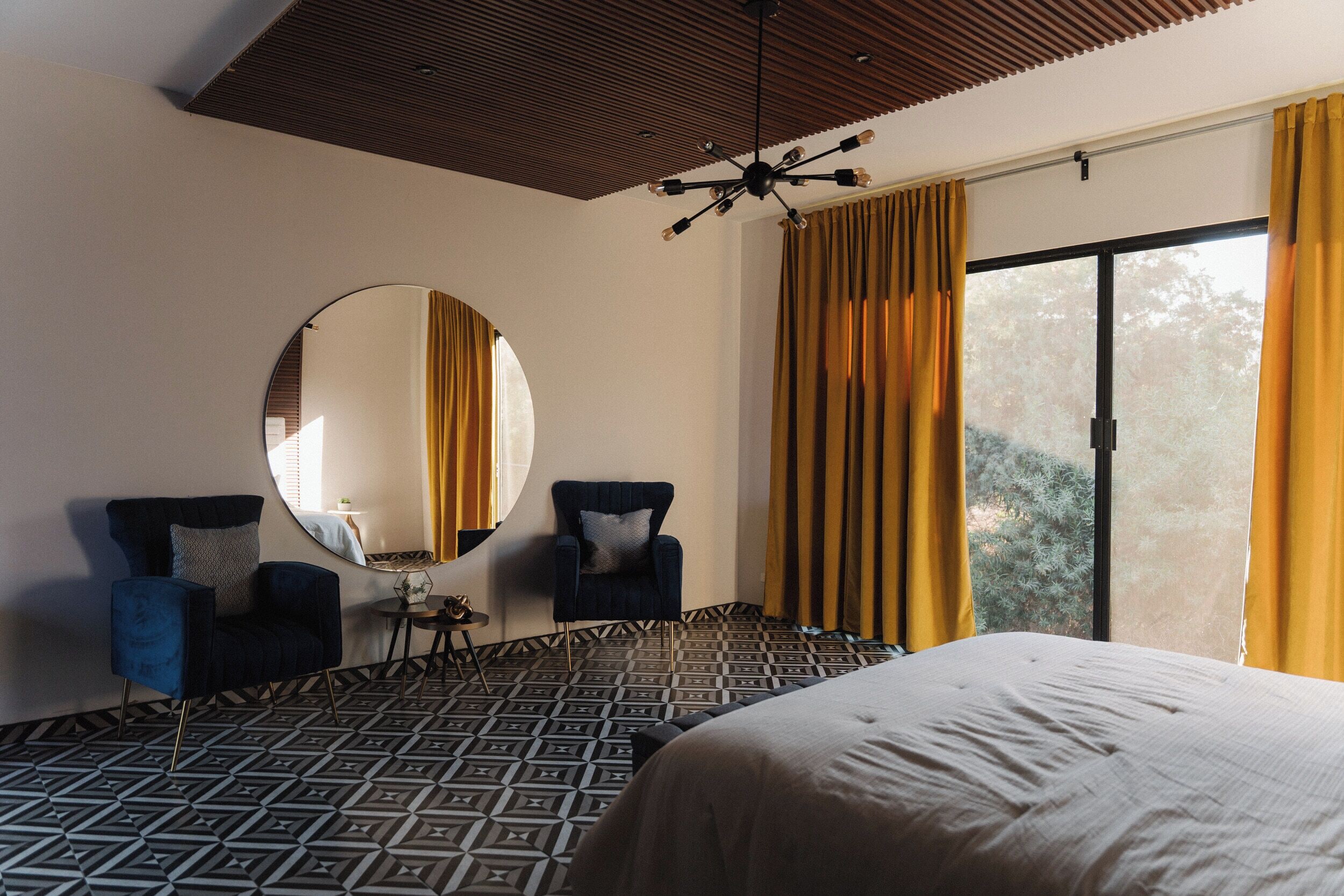 Ein Hotelzimmer mit einem großen Bett, zwei Sesseln, einem runden Spiegel und Blick auf Bäume durch ein großes Fenster.