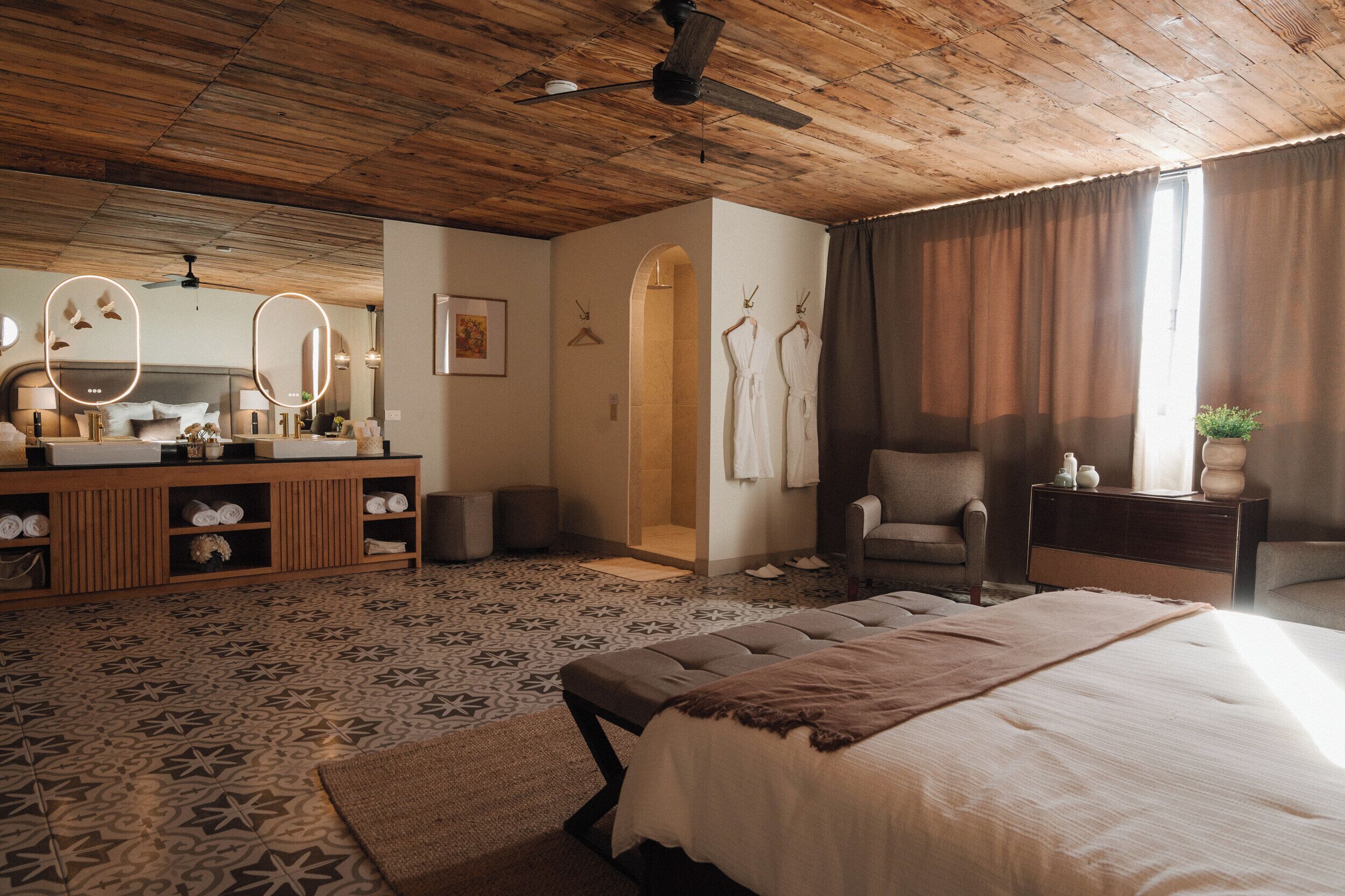 A bedroom with a large bed, a wooden ceiling, a vanity with a mirror, and a seating area with a chair and a small table.