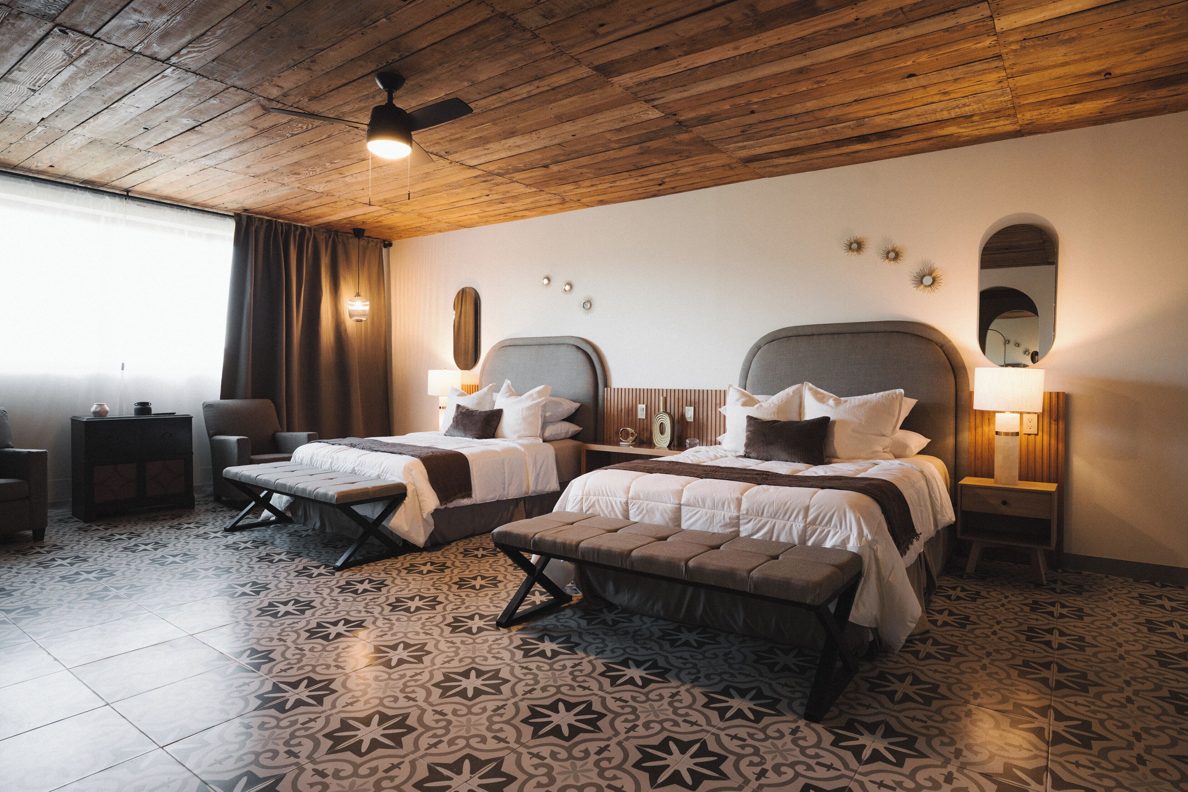 A hotel room with two beds, a ceiling fan, patterned floor tiles, and a window with curtains.