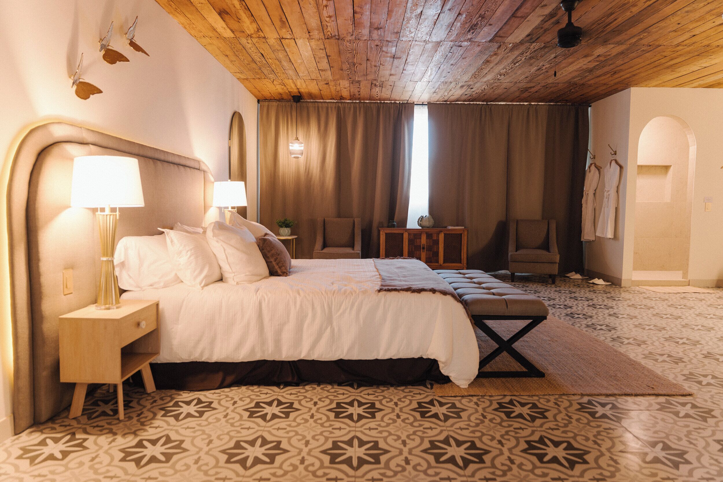 A hotel room with a wooden ceiling, a large bed with white bedding, a bedside table with a lamp, a chair, and a small table with a plant.