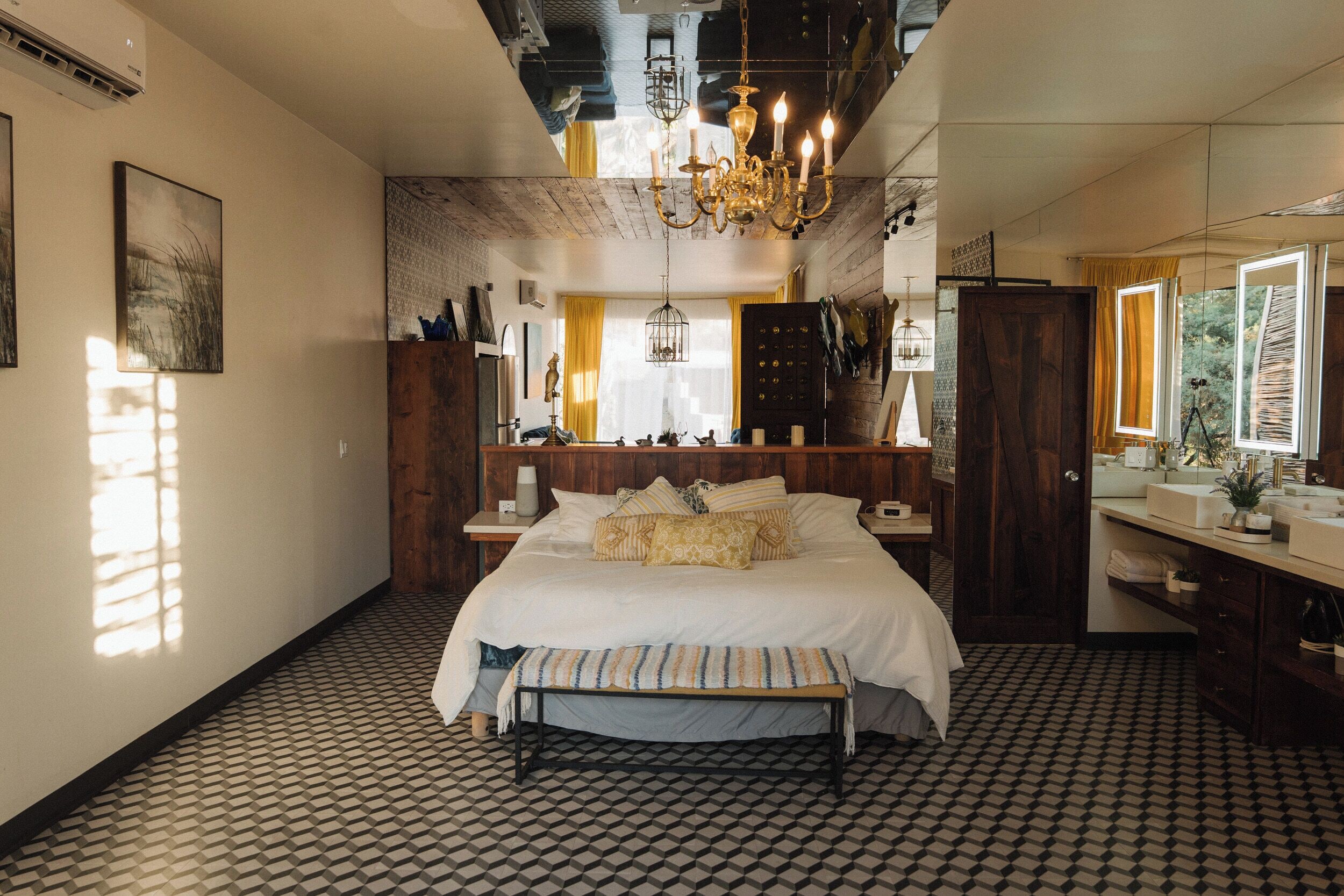A bedroom with a large bed, a wooden headboard, a bench, a chandelier, and a bathroom with a mirror and sink.