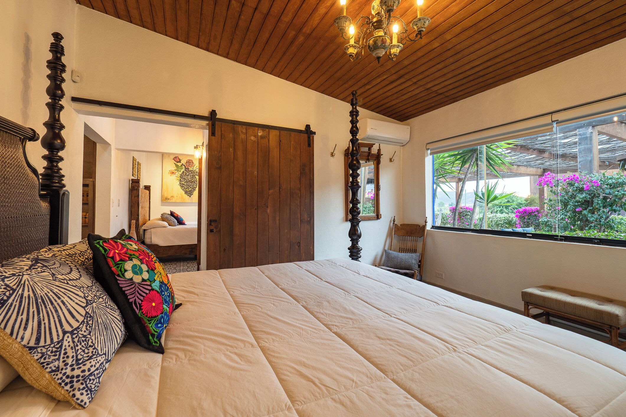 A bedroom with a large bed, a wooden ceiling, a chandelier, a window with a view of greenery, and a door leading to another room.