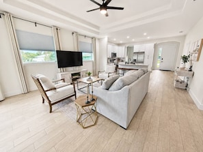 A modern living room with a sofa, armchair, and coffee table. A television is mounted on the wall, and a ceiling fan is visible.
