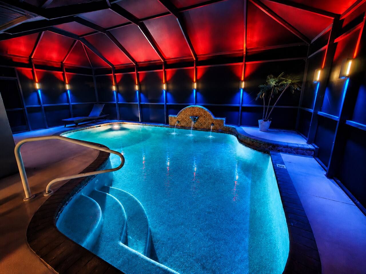 An indoor swimming pool with a geometric ceiling structure, a water fountain, and a poolside area with a metal ladder.