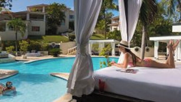 A pool area with a person lying on a bed, another person swimming, and a fountain.