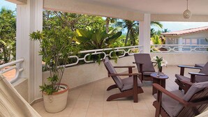 A covered outdoor seating area with chairs and a table, surrounded by potted plants and a view of palm trees and a building.
