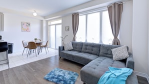 Un salon moderne avec un canapé d’angle gris, un coin repas avec une petite table et des chaises, et de grandes fenêtres avec des rideaux.