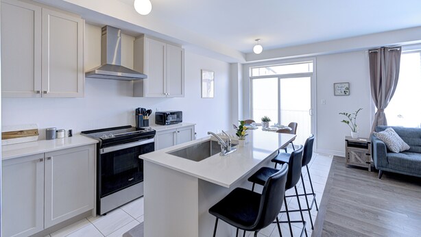 Une cuisine moderne avec des meubles blancs, une cuisinière encastrée et un îlot central doté d’un évier. On y trouve également un coin repas avec une table et des chaises, ainsi qu’un salon avec un canapé.
