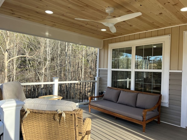 A covered porch with a ceiling fan, a sofa, wicker chairs, and a view of trees.