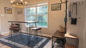 A room with a dining table, two chairs, a chandelier, a window with a view, and a kitchen area with a sink and utensils.