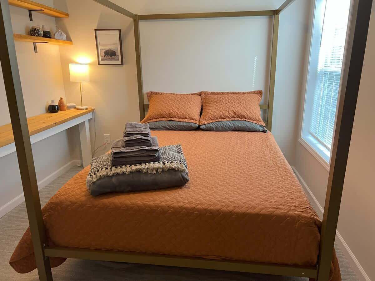 En säng med en sänggavel, ett nattduksbord med en lampa, en inramad tavla på väggen och ett fönster med persienner.