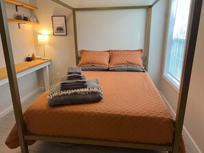 A single bed with a headboard, a bedside table with a lamp, a framed picture on the wall, and a window with blinds.