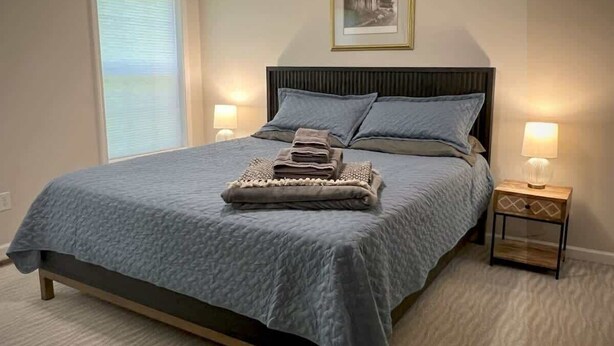 A bedroom with a bed, two bedside lamps, a window with blinds, and a framed picture on the wall.