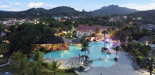 A resort with a large swimming pool, multiple buildings, and a mountainous backdrop.