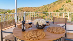 Une table ronde en bois, recouverte de sets de table en fibres tressées, deux chaises en osier, un vase en métal contenant des fleurs blanches, deux verres à vin et une bouteille de vin, le tout placé sur un balcon doté d’une rambarde en métal et offrant une vue sur un paysage vallonné.