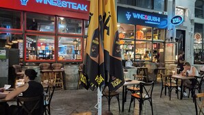 Un espace extérieur aménagé pour les repas, avec des tables et des chaises, un restaurant servant du vin et des sushis en arrière-plan, et un grand parasol.