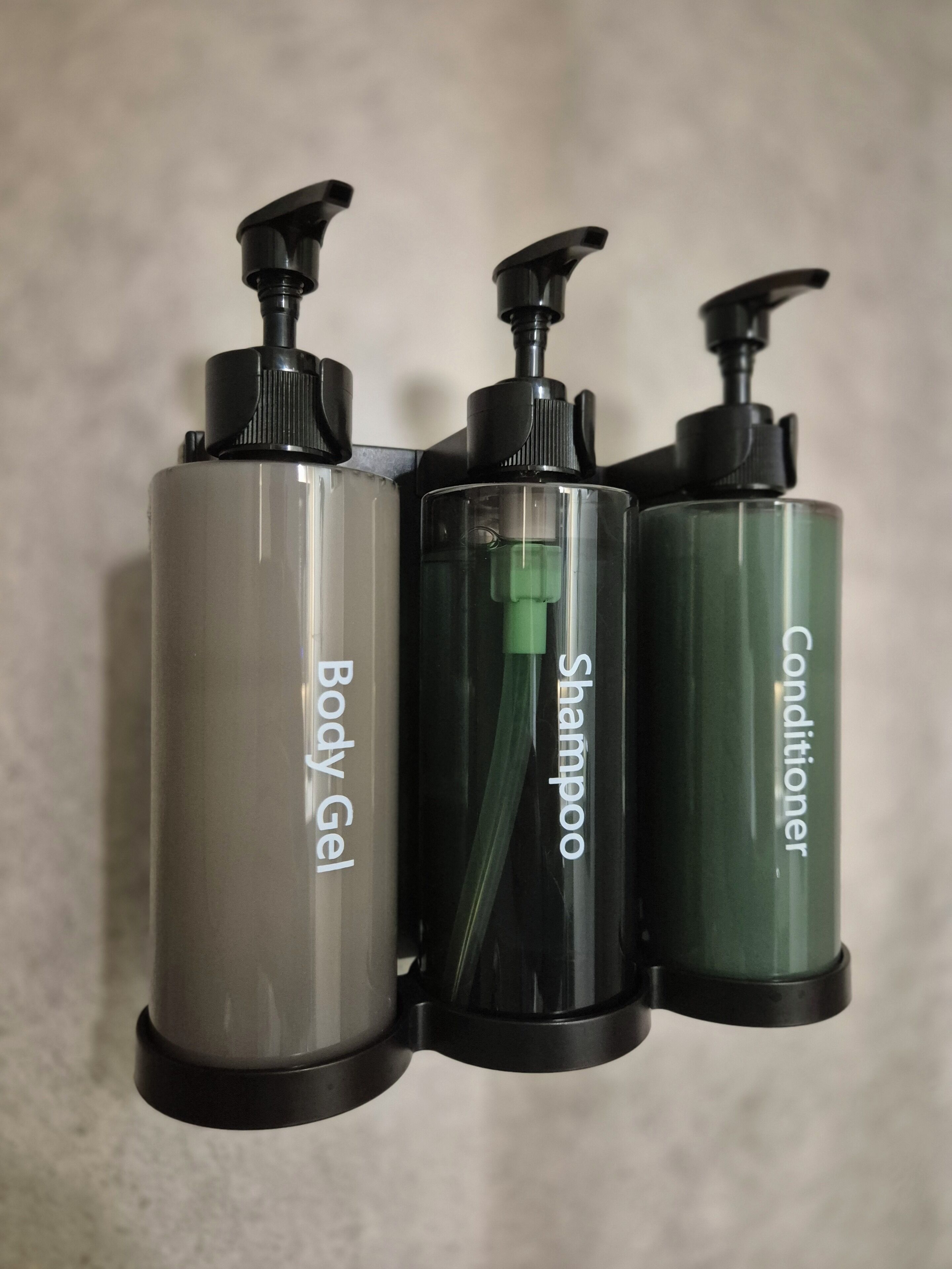 Tres botellas de productos de tocador con dosificadores, etiquetadas como 'Body Gel', 'Champú' y 'Acondicionador'.