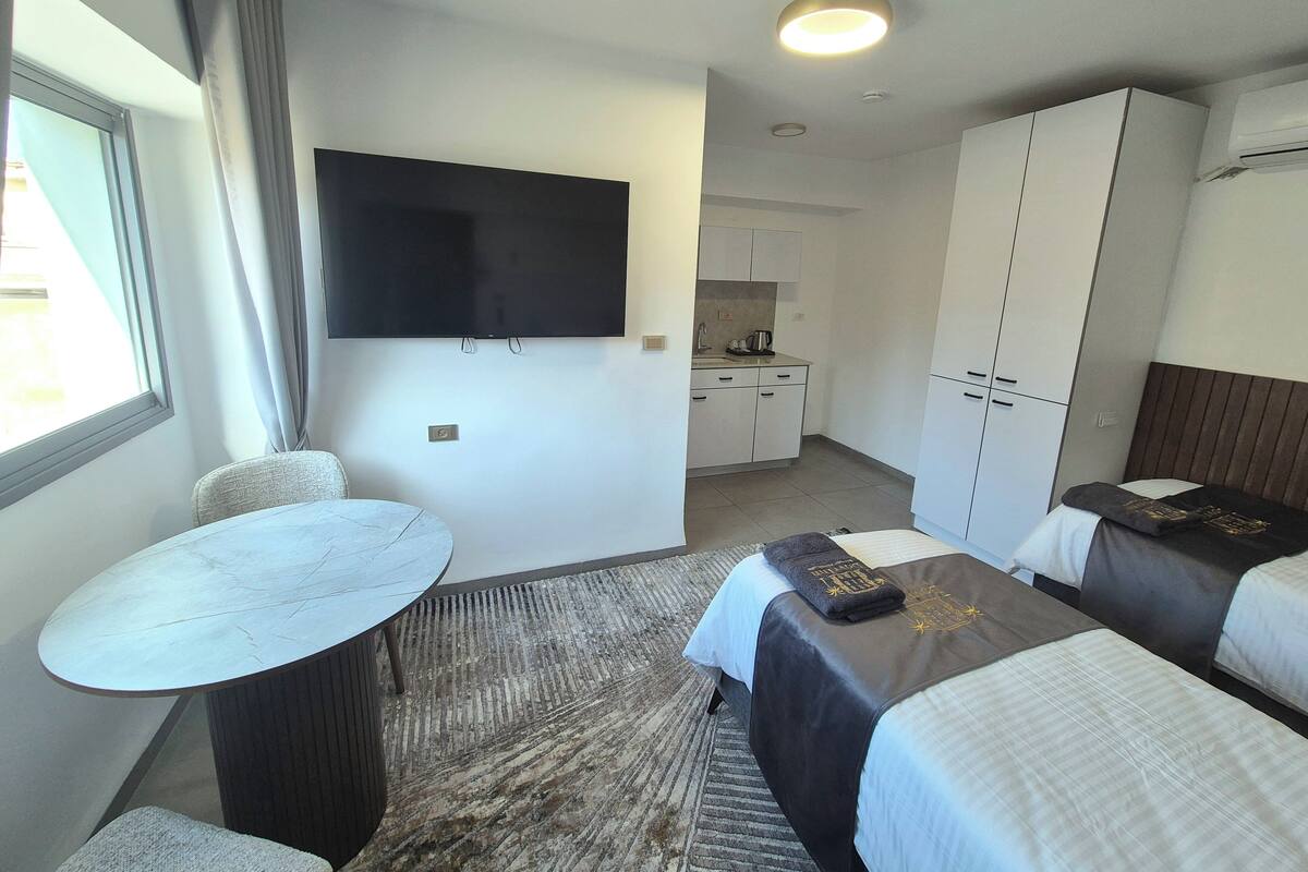 Habitación de hotel con televisión montada en la pared, una pequeña mesa de comedor, dos camas con cabeceros oscuros, kitchenette y un amplio ventanal con cortinas.