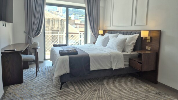 Une chambre d’hôtel équipée d’un lit, d’une table de chevet, d’une lampe, d’une chaise et offrant une vue sur la ville grâce à de grandes fenêtres.
