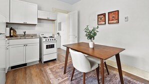 Une cuisine avec des meubles blancs, une cuisinière et une table à manger avec une chaise. Deux tableaux encadrés sont accrochés au mur.