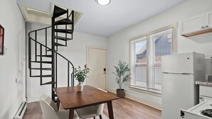 Une cuisine de taille réduite avec un réfrigérateur blanc, un coin repas comprenant une table et des chaises en bois, un escalier en colimaçon noir et une plante en pot.