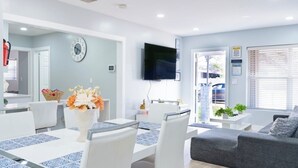 Un salon moderne avec un canapé gris, un coin repas avec une table et des chaises blanches, un téléviseur à écran plat fixé au mur et une fenêtre avec des stores.